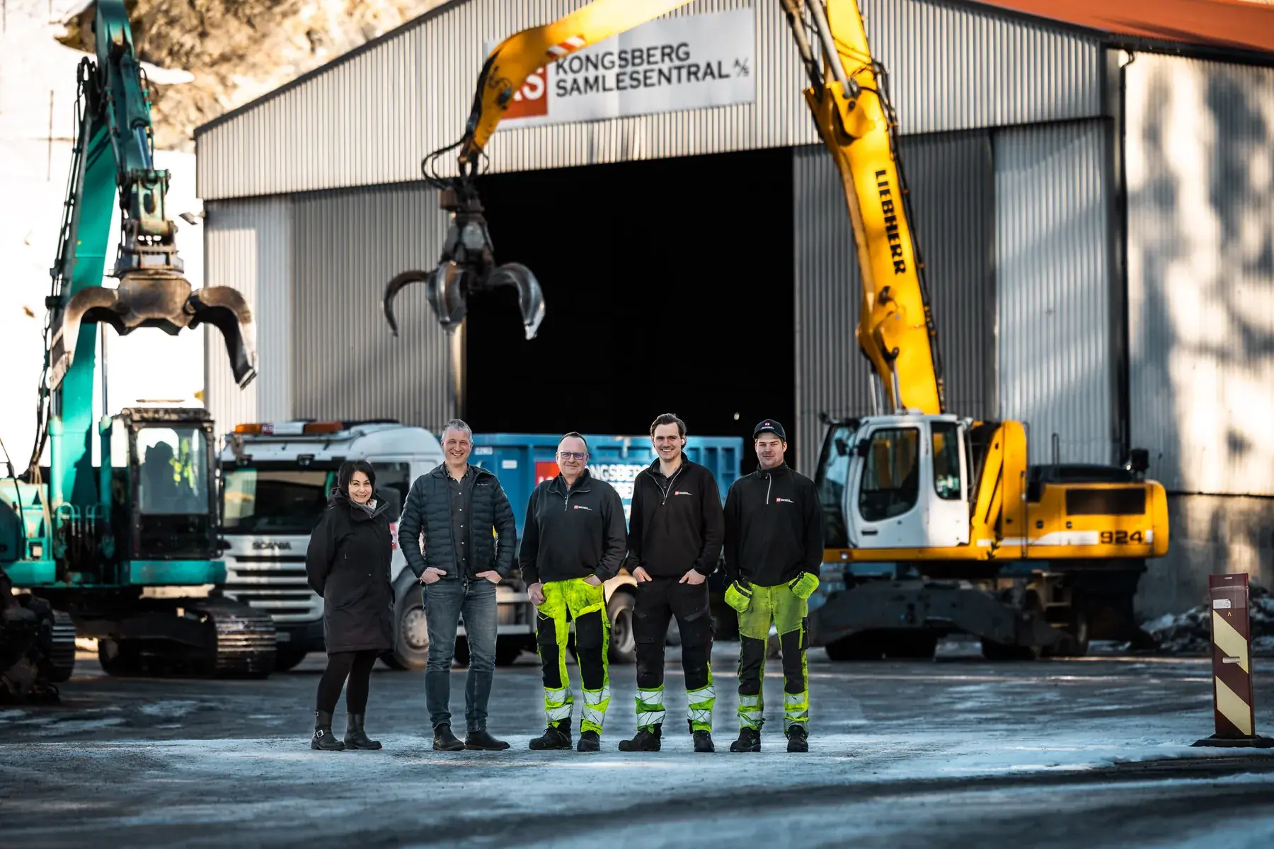 Fem ansatte hos Kongsberg Samlesentral foran hallen med KS-skiltet, gravemaskiner og en blå container på Moane